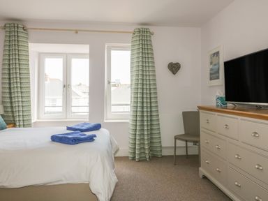 A bedroom with a bed and blue towels next to two windows with patterned curtains a chair and a dresser with a television on top at 5 Ocean Heights in Newquay