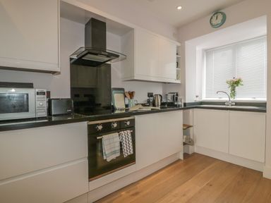 A kitchen with white cabinets black countertops a microwave toaster kettle and a window with blinds at 5 Ocean Heights in Newquay