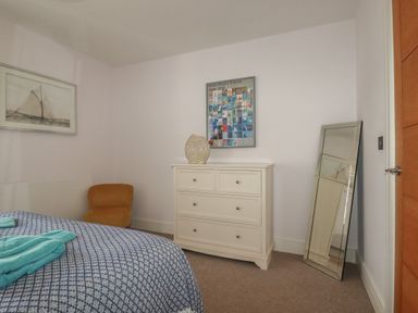A bedroom with a bed covered by a blue and white blanket a brown chair a white chest of drawers a large mirror leaning against the wall and framed pictures on the walls at 5 Ocean Heights in Newquay
