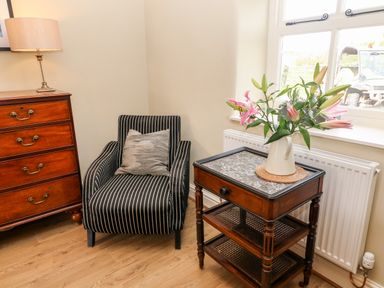 A corner with a striped armchair next to a wooden chest of drawers and a side table with a vase of flowers by a window at Waingate Cottage in Flookburgh