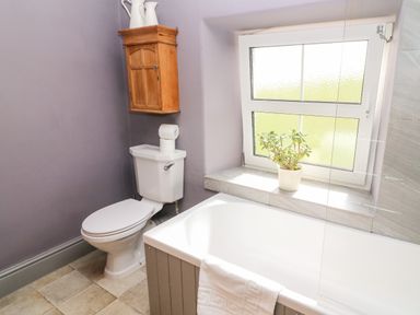 A bathroom with a toilet, bathtub, wooden cabinet, and a potted plant on the windowsill at Waingate Cottage in Flookburgh