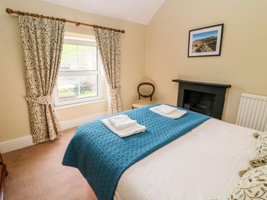 A bedroom with a bed covered with a blue quilt and folded towels near a window with patterned curtains at Waingate Cottage in Flookburgh
