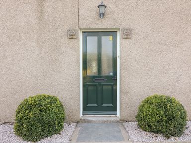 A green front door with frosted glass panels flanked by two bushes at Waingate Cottage in Flookburgh