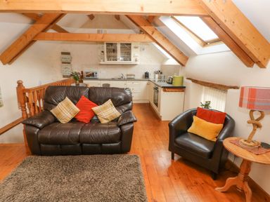 A living area with a brown leather sofa and armchair with cushions a wooden side table with a lamp and a kitchen with white cabinets and wooden beams on the ceiling at Y Cartws in Nevern near Cardigan