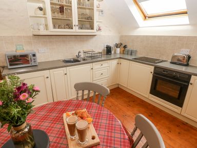A kitchen with white cabinets a countertop with a sink kettle toaster and microwave a table with a red checkered tablecloth and drinks and pastries on a wooden board at Y Cartws in Nevern near Cardigan