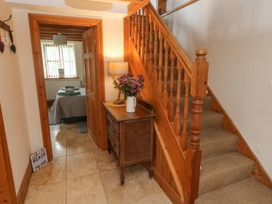 A hallway with a wooden staircase a wooden chest of drawers with a lamp and flowers and a bedroom visible through a doorway at Y Cartws in Nevern near Cardigan