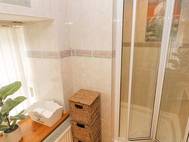 A bathroom corner with a glass shower enclosure a wicker storage unit a white container labeled salts and soaps and a potted plant on a wooden windowsill at Y Cartws in Nevern near Cardigan