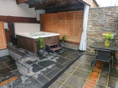 An outdoor patio area with a hot tub two potted plants a metal table with flowers on it and two wicker chairs at Y Cartws in Nevern near Cardigan