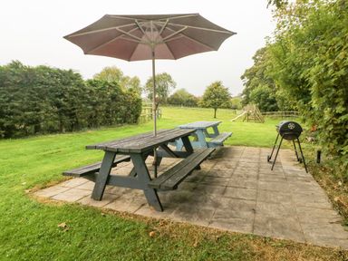 An outdoor patio area with wooden picnic tables and an umbrella next to a barbecue grill with a grassy yard and playground in the background at Y Cartws in Nevern near Cardigan