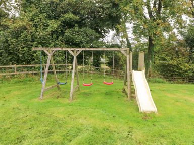 A wooden playground set with three swings a slide and a climbing rope on grass at Y Cartws in Nevern near Cardigan