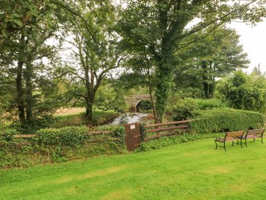 A grassy area with wooden benches and a small gate leading to a stream with a stone bridge surrounded by trees at Y Cartws in Nevern near Cardigan