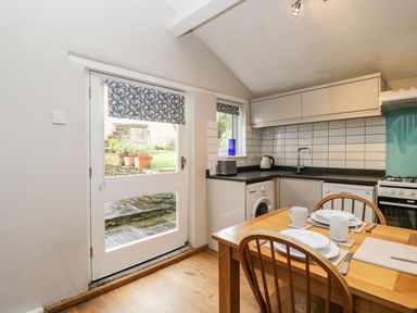 A kitchen with a wooden dining table set for two next to a door and window with patterned blinds at Bower Coach House in Bower Hinton near Martock