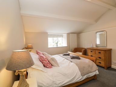 A bedroom with a wooden bed with pillows and blankets a bedside table with a lamp a window a wicker chair and a wooden dresser with a mirror at Bower Coach House in Bower Hinton near Martock