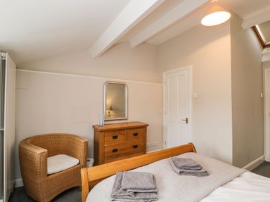 A bedroom with a wooden bed, towels on the bed, a wooden dresser with a mirror above it, a wicker chair, and a closed white door at Bower Coach House in Bower Hinton near Martock