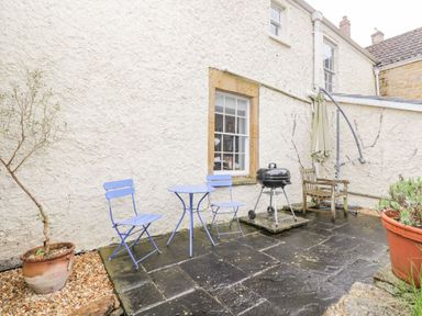 An outdoor patio area with two blue metal chairs and a table a black barbecue grill a wooden chair an umbrella and potted plants at Bower Coach House in Bower Hinton near Martock