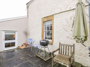A small outdoor patio with a wooden bench charcoal grill blue metal table and chairs and a closed umbrella at Bower Coach House in Bower Hinton near Martock