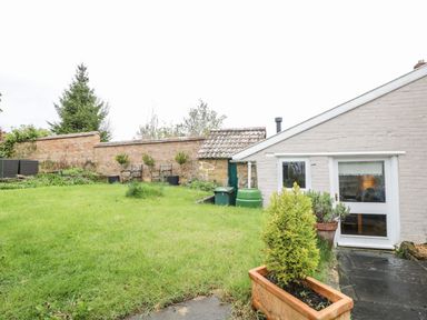 A garden with grass a brick wall wooden chairs and a small house with potted plants at Bower Coach House in Bower Hinton near Martock