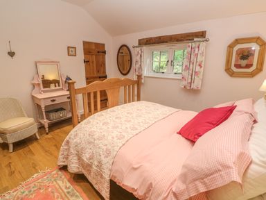 A bedroom with a wooden bed a pink and white bedspread a pink vanity table a window with floral curtains an armchair a wooden door and a framed picture on the wall at Ros Vale in St Buryan