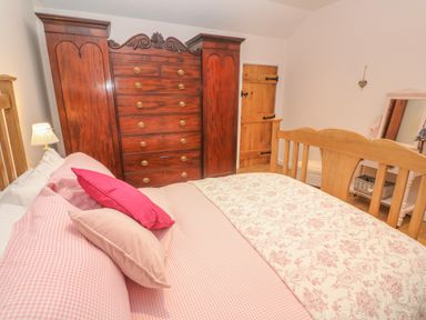 A bedroom with a wooden bed, pink pillows, patterned bedding, a large wooden wardrobe, a wooden door, a pink dressing table with a mirror, and a lamp at Ros Vale in St Buryan