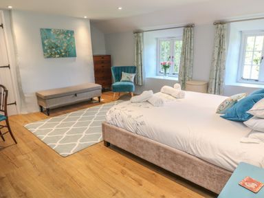 A bedroom with a bed with pillows and towels a chair a bench a chest of drawers and two windows with curtains at Ros Vale in St Buryan