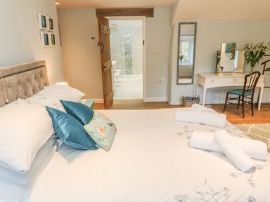 A bedroom with a bed with pillows and rolled towels a dressing table with a chair and mirror and an open door to a bathroom at Ros Vale in St Buryan