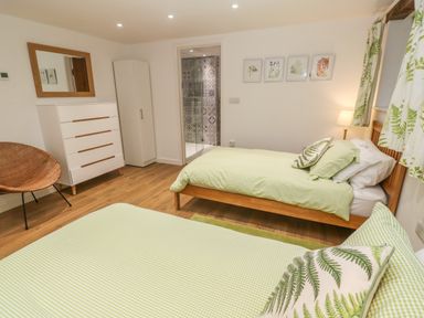 A bedroom with two single beds green check bedding green leaf pillows a white chest of drawers a wicker chair and a bathroom with patterned tiles at Ros Vale in St Buryan