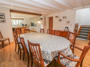 A dining room with wooden chairs around a table covered with a patterned tablecloth near a kitchen and a staircase at Ros Vale in St Buryan