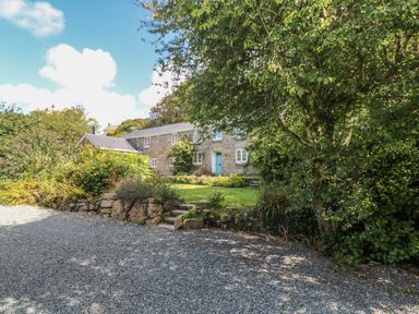 A stone house with a blue door behind stone steps and gravel driveway with trees and bushes at Ros Vale in St Buryan