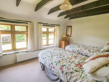 A bedroom with two single beds with floral bedding a wooden chest of drawers a lamp and windows showing greenery at Meadow Lea in Pensilva near Liskeard