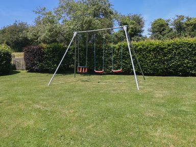 A swingset with three swings on a grassy lawn surrounded by bushes and trees at Meadow Lea in Pensilva near Liskeard
