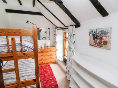 A small bedroom with wooden bunk beds a wooden dresser a window with patterned curtains a red rug a white bookshelf and a painting on the wall at Meadow Lea in Pensilva near Liskeard