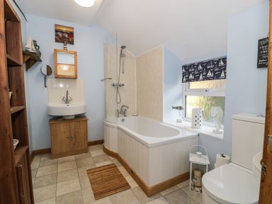 A bathroom with a bathtub shower combination a sink with a wooden cabinet a toilet and a shelf with decorations at 60 Hyfrydle Road Talysarn near Caernarfon