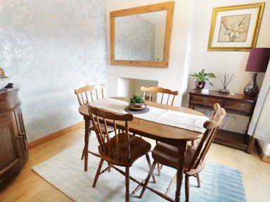 A dining area with a wooden table and four chairs a plant on the table a wooden sideboard with decor a lamp and framed artwork on the wall at 60 Hyfrydle Road in Talysarn near Caernarfon