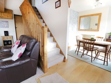 A living room with a brown leather sofa and wooden stairs next to a dining area with a wooden table and chairs at 60 Hyfrydle Road Talysarn near Caernarfon