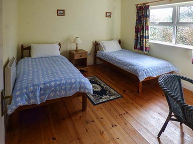 A bedroom with two single beds blue bedspreads a wooden nightstand with a lamp a wicker chair a small rug and a window with curtains at Number 12 in Mulranny County Mayo