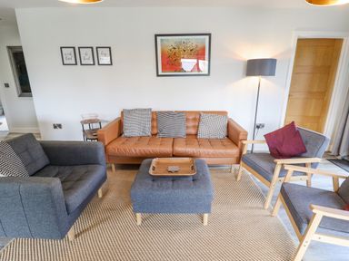 A living room with a brown leather sofa, two gray armchairs, and a gray ottoman with a wooden tray at Lily-pad Lodge in Thorpe-on-the-Hill