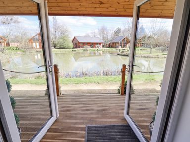 A wooden deck with open glass doors overlooking a pond and wooden cabins at Lily-pad Lodge in Thorpe-on-the-Hill