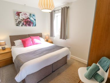A bedroom with a bed with pink pillows and a gray throw a chair with green towels and a painting of flowers on the wall at Lily-pad Lodge in Thorpe-on-the-Hill