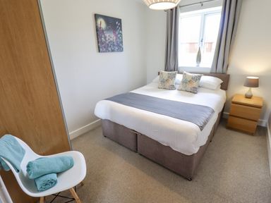 A bedroom with a double bed two pillows a bedside table with a lamp a window with curtains a painting on the wall a chair with towels at Lily-pad Lodge in Thorpe-on-the-Hill