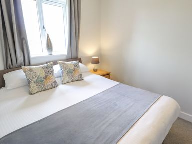 A bedroom with a bed two floral pillows a gray runner a bedside table with a lamp and a window at Lily-pad Lodge in Thorpe-on-the-Hill