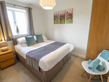 A bedroom with a double bed two blue cushions a bedside table with a lamp a window with curtains and a chair with towels at Lily-pad Lodge in Thorpe-on-the-Hill