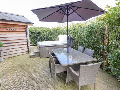 An outdoor wooden deck with a glass table, six wicker chairs, a large umbrella, a hot tub, and greenery at Lily-pad Lodge in Thorpe-on-the-Hill