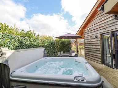 An outdoor patio area with a hot tub a table with chairs and an umbrella next to a wooden cabin at Lily-pad Lodge in Thorpe-on-the-Hill