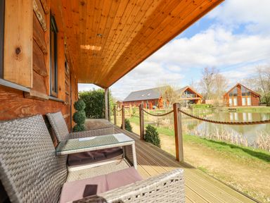 A wooden porch with wicker furniture overlooking a pond and wooden lodges at Lily-pad Lodge in Thorpe-on-the-Hill