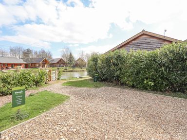 A gravel parking area beside a wooden lodge with a pond and more lodges in the background at Lily-pad Lodge in Thorpe-on-the-Hill