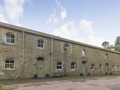 A stone building with multiple green doors and windows along a paved road at Gardeners Cottage in Bellingham