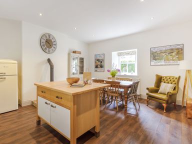 A kitchen and dining area with a wooden island, dining table with chairs, armchair, refrigerator, and wall clock at Gardeners Cottage in Bellingham