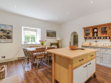 A kitchen dining area with a wooden dining table and chairs a kitchen island and shelves with cups at Gardeners Cottage in Bellingham