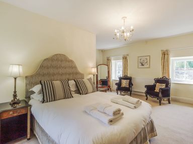 A bedroom with a large bed with striped pillows two armchairs a floor mirror and two windows at Gardeners Cottage in Bellingham