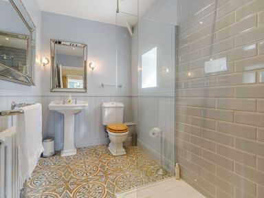A bathroom with a pedestal sink, toilet with wooden seat, patterned tile floor, glass shower enclosure and tiled shower wall at Gardeners Cottage in Bellingham